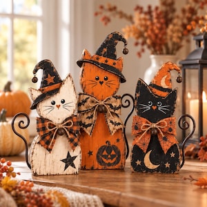 以下が含まれることがあります： ハロウィンの装飾用の木製猫の置物3体。左の猫は白で黒い星とチェック柄のリボン付き。中央の猫はオレンジ色でカボチャとコウモリ柄のリボン付き。右の猫は黒で三日月とクモの巣柄のリボンが付いています。各猫は魔女の帽子をかぶり、金属製の尾が付いています。