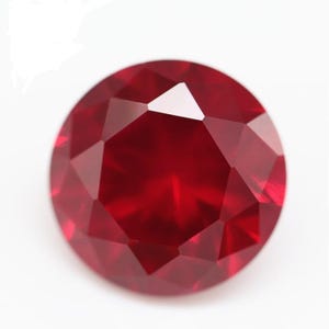 Puede incluir: Una piedra preciosa redonda y facetada de un color rojo intenso y vibrante. La piedra presenta un corte brillante con múltiples facetas triangulares y en forma de cometa que reflejan la luz, creando una superficie clara y pulida.