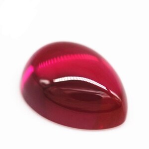 May include: A pear-shaped cabochon gemstone with a deep, vibrant red color and a smooth, polished surface. The stone features a rounded dome top and a flat base, reflecting light with a glossy sheen across its translucent, saturated surface.