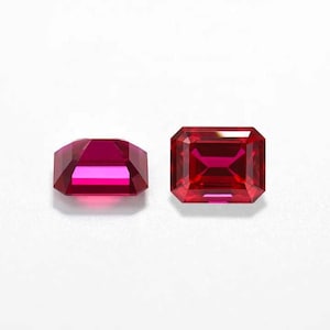 May include: Two vibrant red gemstones with an emerald cut, showcasing a rectangular shape and faceted surfaces. One stone is positioned at a slight angle to display its depth, while the other is viewed directly from the top, highlighting its clarity.