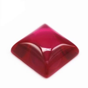 May include: A square-shaped, deep red gemstone with a smooth, polished surface and a faceted pyramid-like top. The translucent material displays a rich crimson hue with subtle light reflections on its curved edges and a flat base.