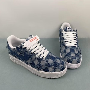 Puede incluir: Un par de zapatillas de corte bajo con una parte superior de mezclilla a cuadros azules y blancos con bordes deshilachados. Los zapatos tienen cordones blancos, una entresuela blanca y un forro interior blanco. Una correa blanca con texto naranja está unida a la lengüeta.