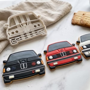 Puede incluir: Un cortador de galletas blanco con forma de coche junto a tres galletas decoradas con la parte delantera de un coche. Las galletas están glaseadas en negro, rojo y blanco, con detalles de rejillas, faros y parachoques. Dos galletas simples son visibles al fondo.
