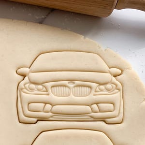 May include: A beige cookie dough sheet featuring a pressed impression of the front view of a car with a distinct dual-kidney grille, headlights, and bumper design. A wooden rolling pin is positioned at the top edge of the dough surface.