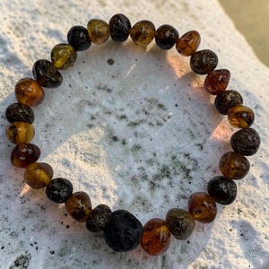 Può includere: Un braccialetto elastico composto da perline irregolari e arrotondate in varie tonalità di miele traslucido, ambra profonda e marrone scuro opaco. Le perline presentano texture naturali e sfumature di colore disposte in cerchio.