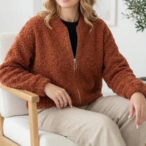 May include: A rust-colored, long-sleeved bomber jacket made of a textured, fuzzy fleece material. The jacket features a full-length silver-toned metal zipper closure down the center front, a simple round neckline, and ribbed cuffs at the wrists.