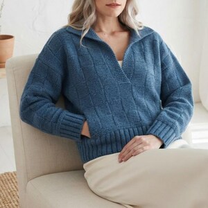 May include: A blue long-sleeve sweater featuring a textured checkerboard knit pattern, a wide collar, and a deep V-neckline. The garment includes ribbed cuffs and a ribbed hem, along with a front pocket opening where a hand is tucked inside.