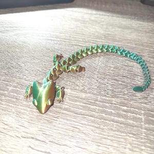 May include: A 3D printed articulated lizard figurine featuring a colour-shifting iridescent finish that transitions between metallic green and gold tones. The lizard has a segmented body, a long curved tail, and four small legs with visible joints.