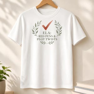 May include: A white short-sleeve t-shirt featuring a graphic of a red checkmark above a green leaf wreath. The text inside the wreath reads ELA: RED PENS & PLOT TWISTS in black capital letters. The shirt is displayed on a wooden hanger.