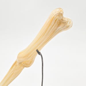 May include: A decorative bone-shaped object with a light yellow and white swirled pattern. The bone is supported by a thin, black metal stand and rests on a gray, textured surface. The background is plain white.
