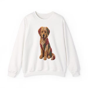 May include: A white crewneck sweatshirt featuring a central graphic of a golden-colored dog. The dog is depicted with a patchwork-style coat in shades of tan, red, and green, decorated with small heart and star patterns throughout its fur.