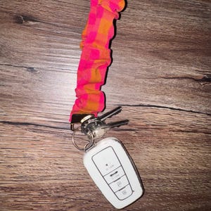 May include: A fabric keychain wristlet featuring a bright pink and orange checkered pattern. The wristlet is attached to a gold-toned metal clasp holding a silver metal key ring with two metal keys and a white rectangular car key fob.