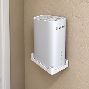 May include: A white rectangular Gfiber router mounted on a wall using a white plastic bracket. The device features a top mesh vent, a multi-colored logo, and four vertical status indicator lights with icons for power, internet, and Wi-Fi connectivity.