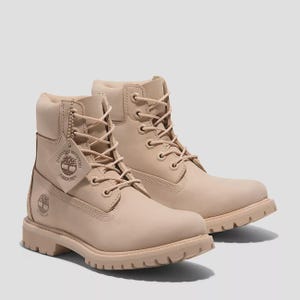 Puede incluir: Un par de botas impermeables de color beige claro con cordones, ojales metálicos, cuello acolchado y suela gruesa con tacos. Una etiqueta cuadrada unida a los cordones muestra el logotipo del árbol de Timberland y el texto Guaranteed Waterproof.