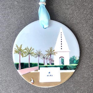 May include: Round ceramic ornament with a light blue background, featuring a painted scene of a white tower, palm trees, and a small figure. The ornament is suspended by a light blue ribbon and a silver ring. The word "ALYS" is visible.
