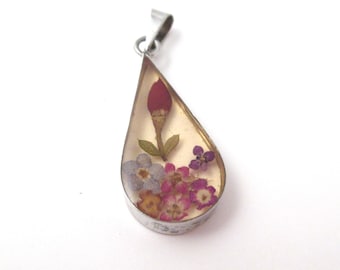 May include: A teardrop-shaped pendant featuring a silver-toned metal frame filled with clear resin. Inside the resin are preserved pressed flowers in shades of pink, purple, and blue, along with a single red flower bud and green leaves.