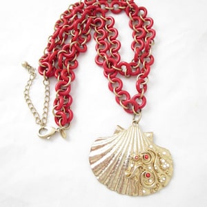 May include: A gold-colored seashell pendant necklace with a red chain. The seashell pendant is adorned with red and white embellishments. The necklace has a lobster clasp closure.
