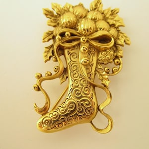 May include: A gold-colored brooch in the shape of a Christmas stocking, adorned with a bow, holly leaves, and berries. The stocking has an ornate, swirling design. The brooch is likely made of metal and is a festive accessory.