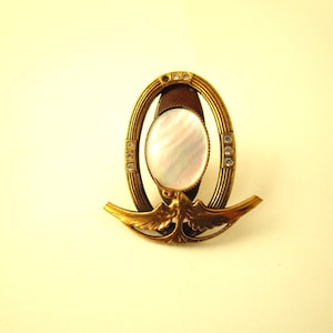 May include: Vintage gold-tone brooch with an oval mother-of-pearl center. The brooch features a decorative frame with small clear crystals and a stylized bird design at the base. The brooch has a classic, elegant design.