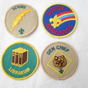 May include: Four embroidered patches with various designs. One patch features a gold quill and the word "SCRIBE". Another has a fleur-de-lis and the words "DONOR AWARENESS". A third patch says "LIBRARIAN" and depicts books. The last patch says "DEN CHIEF" with a tiger.