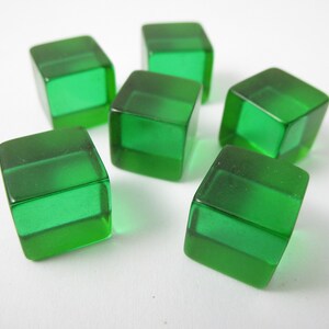 May include: Six translucent green dice are scattered on a white surface. Each die is a cube shape with rounded edges. The dice are a vibrant, emerald green color, and appear to be made of a solid, clear material. The image is well-lit, highlighting the dice's details.