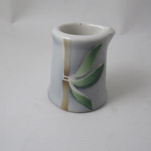 May include: A small, white ceramic creamer with a blue and green bamboo design. The creamer has a rounded base and a slightly flared top.