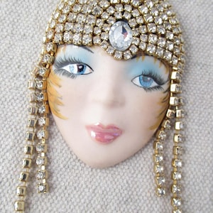 May include: A decorative face brooch with a gold-tone headpiece and dangling strands, adorned with clear rhinestones. The face has blue eyes, pink lips, and a painted complexion. A large, clear rhinestone is centered on the headpiece.