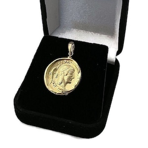 May include: A gold-colored pendant featuring a profile of a person in a circular design. The pendant is displayed inside a black velvet jewelry box with a white satin interior. The pendant has a small loop at the top for a chain.