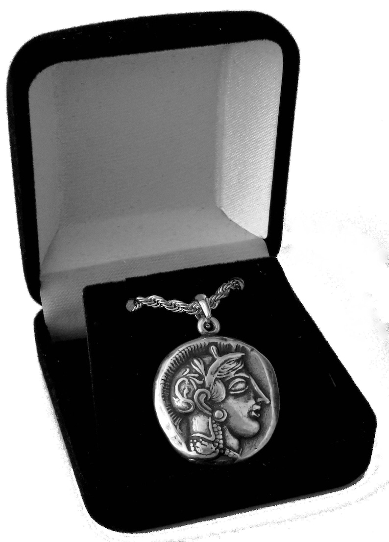 May include: A silver pendant necklace featuring a detailed profile of a classical figure, presented in a black velvet jewelry box. The pendant is round and has intricate detailing. The chain is a twisted rope style.