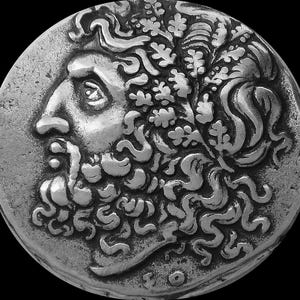 May include: Silver coin with a detailed profile of a man with a beard and a wreath of leaves on his head. The coin is in good condition and has a smooth surface.