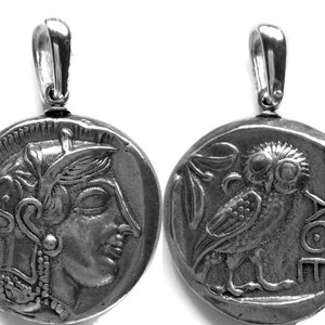 May include: Silver-toned pendant featuring a detailed replica of an ancient Greek coin. One side depicts a profile of a woman's face, the other an owl with the inscription "AOE". The pendant has a loop for a chain.