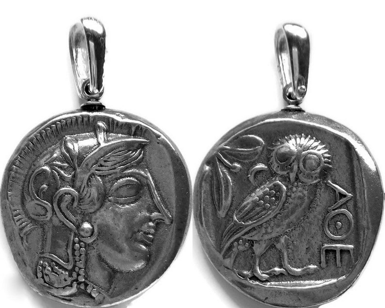 May include: Silver-toned pendant featuring a detailed replica of an ancient Greek coin. One side depicts a profile of a woman's face, the other an owl with the inscription "AOE". The pendant has a loop for a chain.