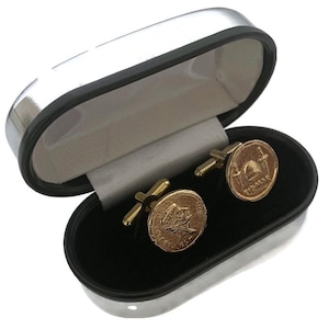 May include: A pair of gold-toned cufflinks with coin-like faces, displayed in an open, silver-trimmed black case. The cufflinks feature detailed designs and are presented on a black velvet-like interior.