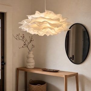 May include: A large, white, cloud-like pendant light fixture with multiple layered, ruffled, and translucent paper-like petals. The light emits a soft, warm glow from within, creating a textured, organic shape that hangs from a single thin cord.
