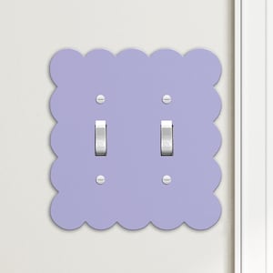 May include: A light purple, scalloped-edge double light switch cover. The cover has two toggle switches and four small screws. The wall is white, and the door frame is visible on the right side.