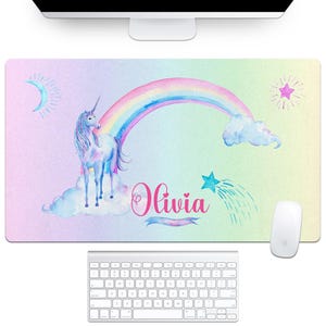 a desk pad with a pastel rainbow background, a unicorn on the left standing on clouds, a rainbow arc across the center and a shooting star on the right.  personalized with a name in a pink cursive font.  from the brand aqua magnolia.