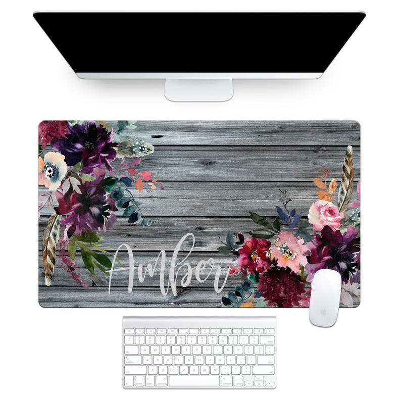a personalized desk pad with a grey wooden plank background and floral design in shades of purple, red, and white along the edges, with gold feathers also incorporated into the design.