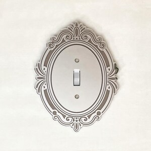 May include: A silver-toned oval light switch plate featuring an intricate, symmetrical filigree border design. The plate is secured with two small screws and includes a central toggle switch with the word ON printed on the lever.