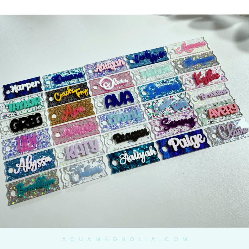 a collection of personalized tumbler name tags and name plates, each with a custom acrylic name on it, each with a different design and color scheme including glitter, confetti, chunky glitter and more. The tags are arranged in rows and columns, showcasing a variety of names and styles.