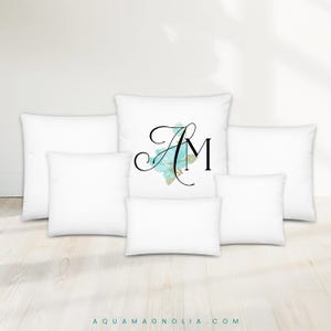 May include: White throw pillows in various sizes. One pillow features the letters "AM" in black script with a blue floral design. The pillows are arranged on a light wood floor. The website address "AQUAMAGNOLIA.COM" is at the bottom.