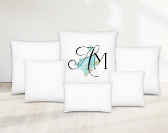 May include: White throw pillows in various sizes. One pillow features the letters "AM" in black script with a blue floral design. The pillows are arranged on a light wood floor. The website address "AQUAMAGNOLIA.COM" is at the bottom.