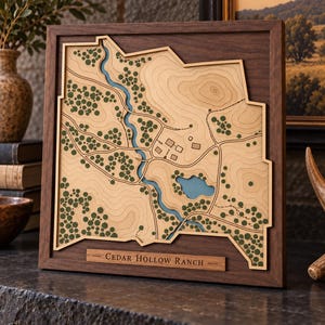 May include: A square wooden wall art piece featuring a layered topographic map of Cedar Hollow Ranch. The map displays a winding blue river, a small blue lake, green circular tree icons, and tan terrain contours, all encased in a dark wood frame.