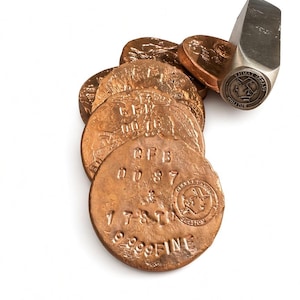 May include: A stack of circular copper-colored metal discs with stamped text including CFB 0087, 17.8 TO, and 999 FINE, alongside a metal stamping tool featuring a circular logo with a profile of a person wearing a cap and the text JIMAT.