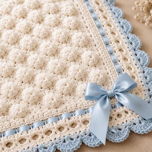 May include: A cream-colored crochet blanket featuring a textured bobble stitch pattern throughout the center. The border is accented with a woven light blue satin ribbon and finished with a light blue scalloped crochet edge and a satin bow.