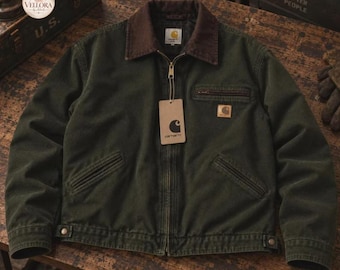 May include: A dark olive green Carhartt jacket featuring a brown corduroy collar, a full-length front zipper, and a chest pocket with a zipper closure. A rectangular Carhartt brand tag is attached to the zipper pull, and a small square logo patch is sewn onto the chest.
