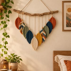 May include: A macrame wall hanging featuring multiple fiber feathers in shades of rust, navy blue, sage green, mustard yellow, and cream, suspended from a natural wooden branch with jute twine and small wooden beads.