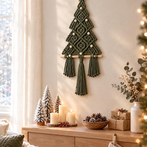 May include: A wall-hanging macrame Christmas tree crafted from dark olive green cord, featuring a wooden star topper, light-colored wooden beads as ornaments, and a central tassel base suspended from a horizontal wooden dowel rod.