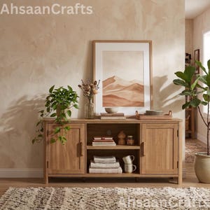 May include: A light wood media console with two side cabinets and an open center shelf. The console holds a potted plant, a framed desert landscape print, books, folded textiles, and mugs. The text AhsaanCrafts appears in the corners.