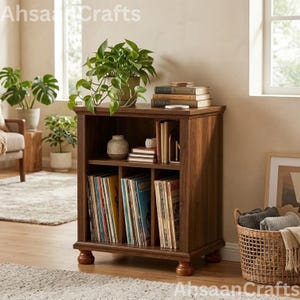 May include: A dark wood record storage cabinet with four bun feet, featuring an open top shelf holding a potted plant, books, and a small vase, and a lower section with three vertical dividers filled with vinyl record albums.