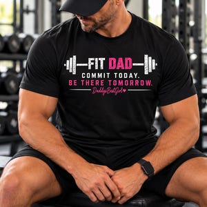 May include: A black short-sleeved t-shirt featuring a white and pink graphic of a barbell with the text FIT DAD, COMMIT TODAY. BE THERE TOMORROW. and DaddysBestGirl with a small heart icon. The shirt has a crew neckline and a standard fit.